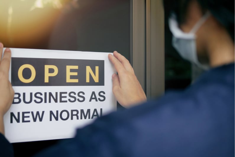 Masked person putting sign in door that read, "OPEN BUSINESS AS NEW NORMAL"